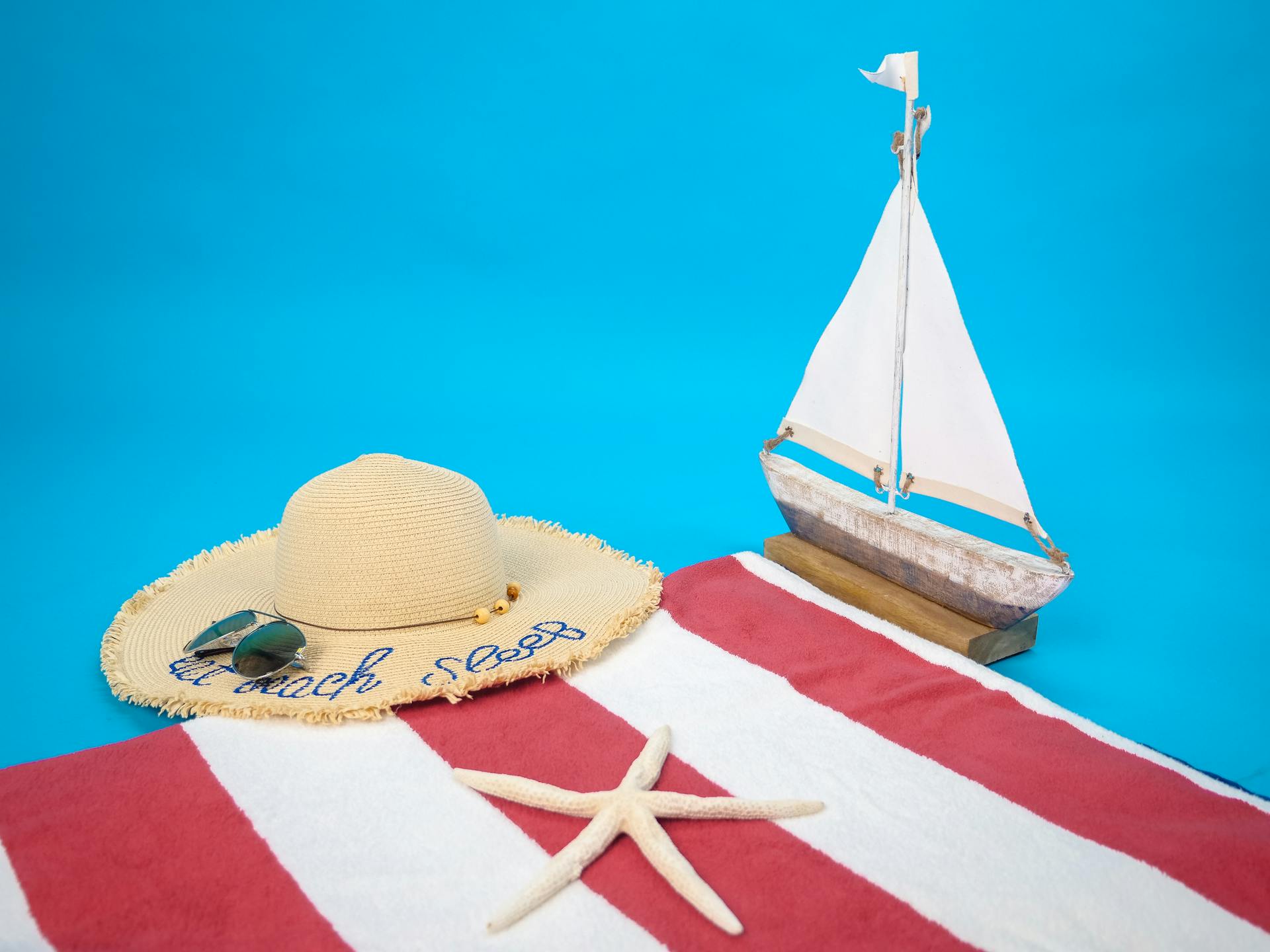 Flat lay of summer beach essentials including sun hat, towel, and model sailboat.