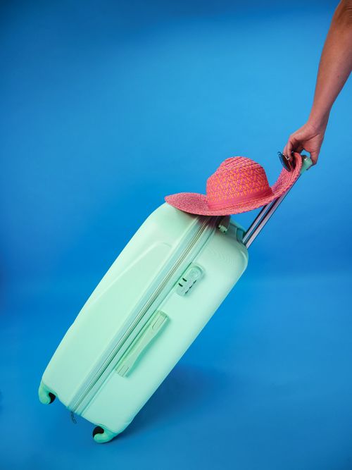 Free Mint green suitcase with pink sun hat against a blue background, evoking summer travel vibes. Stock Photo
