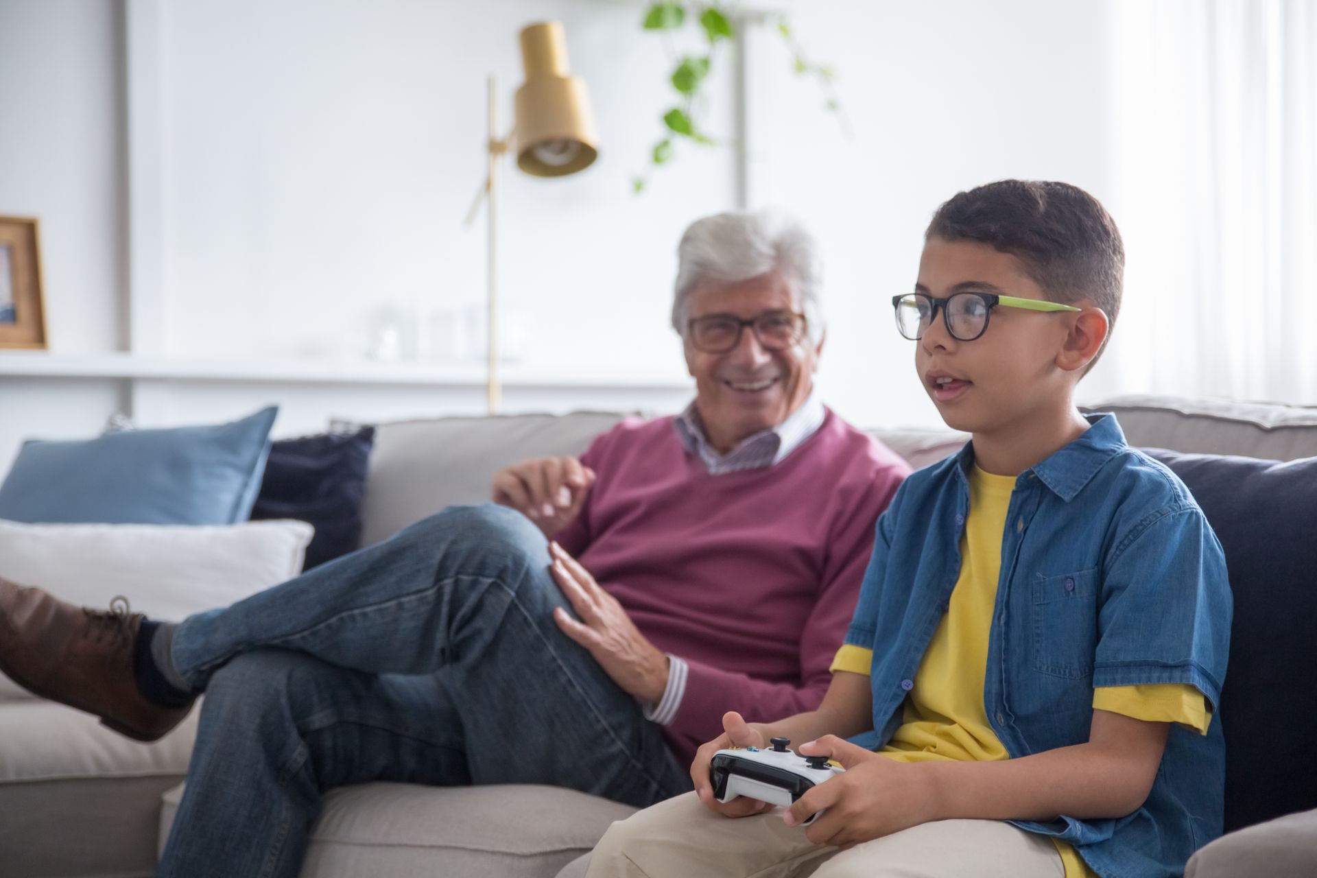 Grandfather and grandson enjoying leisure time indoors, playing video games on a sofa.
