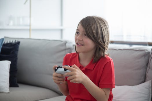 A happy child in a red polo shirt plays video games indoors, sitting on a sofa.