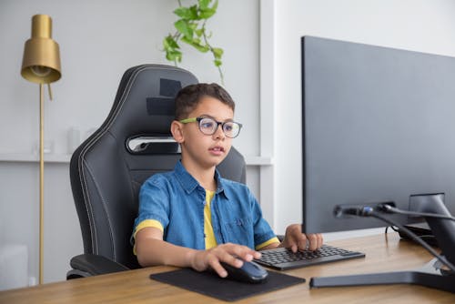 Little Boy Wearing Eyeglasses while Sitting on Gaming Chair in Front of the Computer