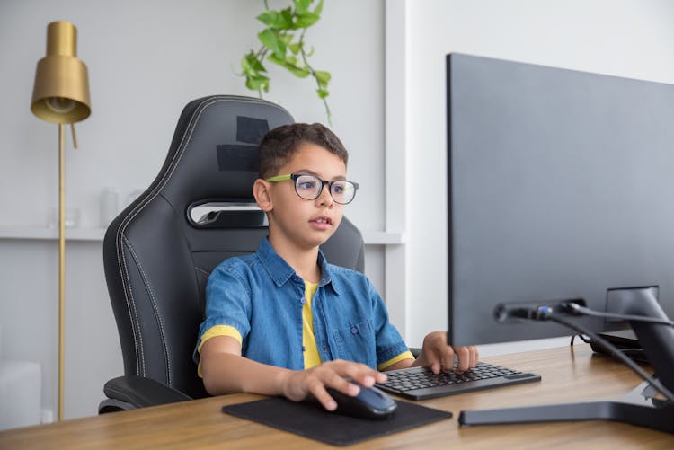 Little Boy Wearing Eyeglasses While Sitting On Gaming Chair In Front Of The Computer