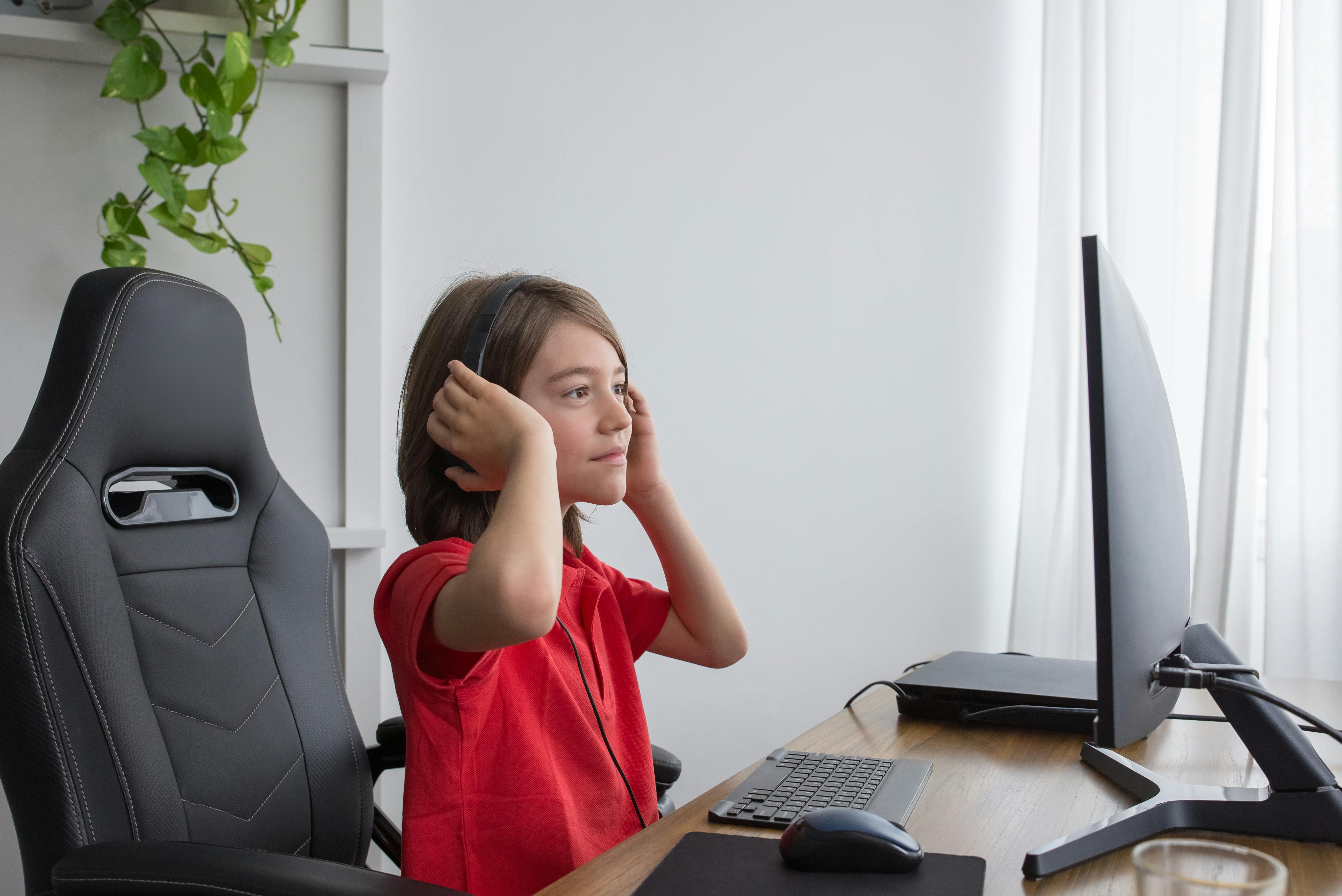 A young boy in a red shirt sits at a computer desk, wearing headphones and enjoying gaming at home.