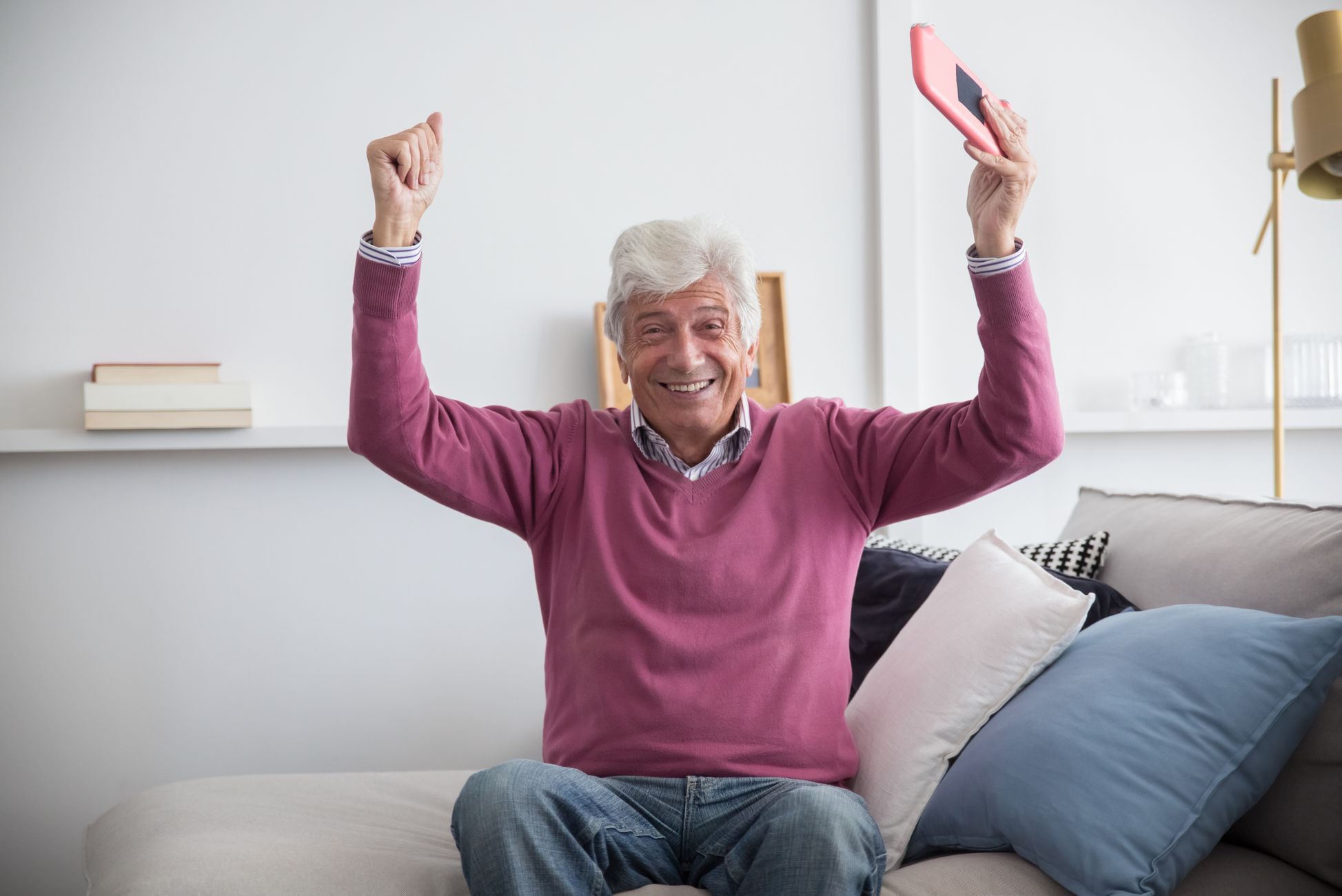 A happy senior man sitting on a sofa holding a smartphone indoors, celebrating with raised arms.
