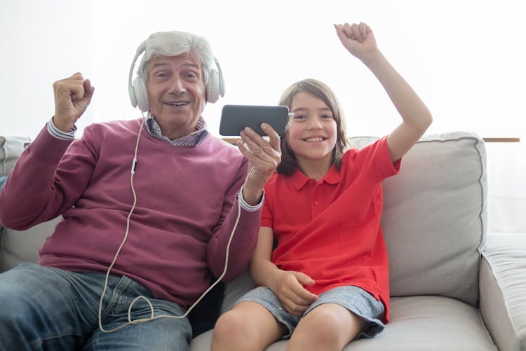 Elderly Man Holding Cellphone With Headset Sitting On Gray Sofa Beside A Girl In Red Polo Shirt 