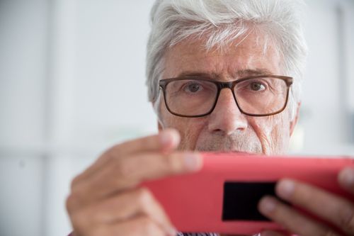 Free Close-up of a senior man with eyeglasses playing with a red handheld gaming console indoors. Stock Photo