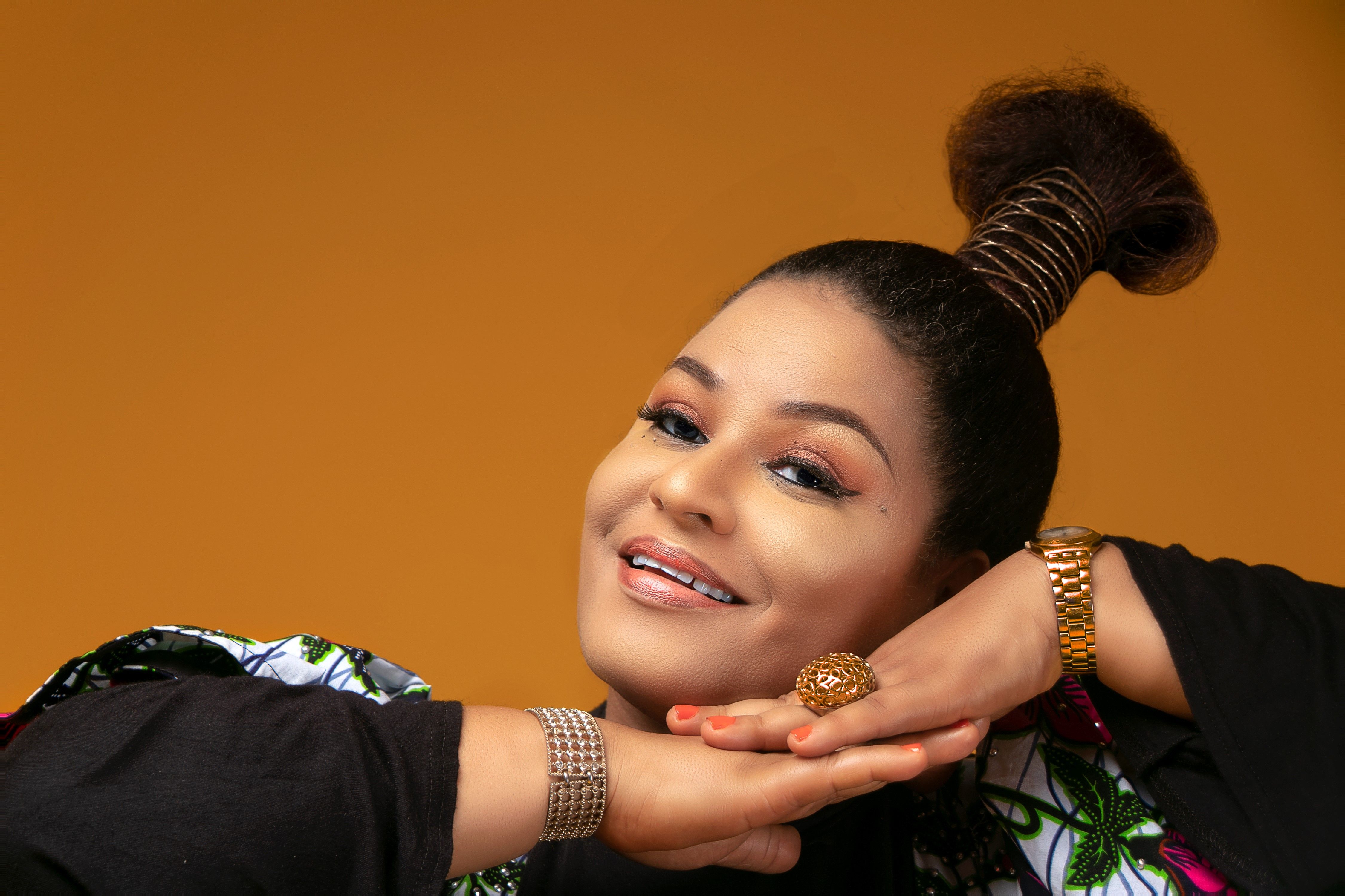 Positive female with makeup with black hair in high bun wearing wristwatch and accessories looking at camera on brown background in studio