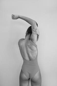 Black and white photo of a woman stretching in a bodysuit, emphasizing form and elegance.