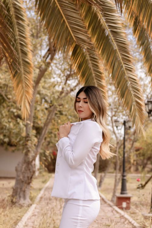 Free A woman gracefully poses under palm trees wearing an elegant white suit, offering a sense of sophistication. Stock Photo