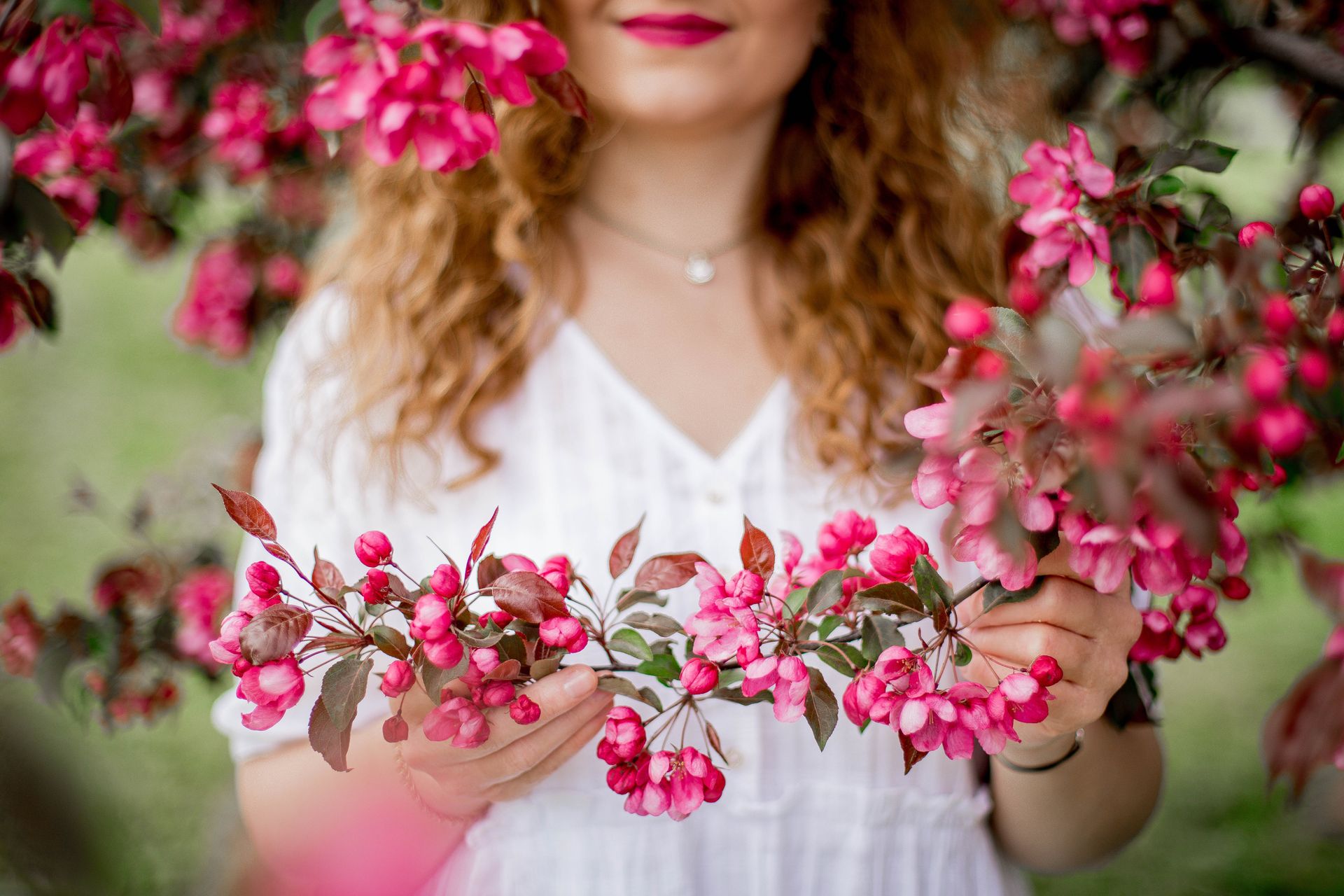 Crop content anonymous female with curly hair touching apple branch with small flowers while standing in garden on blurred background