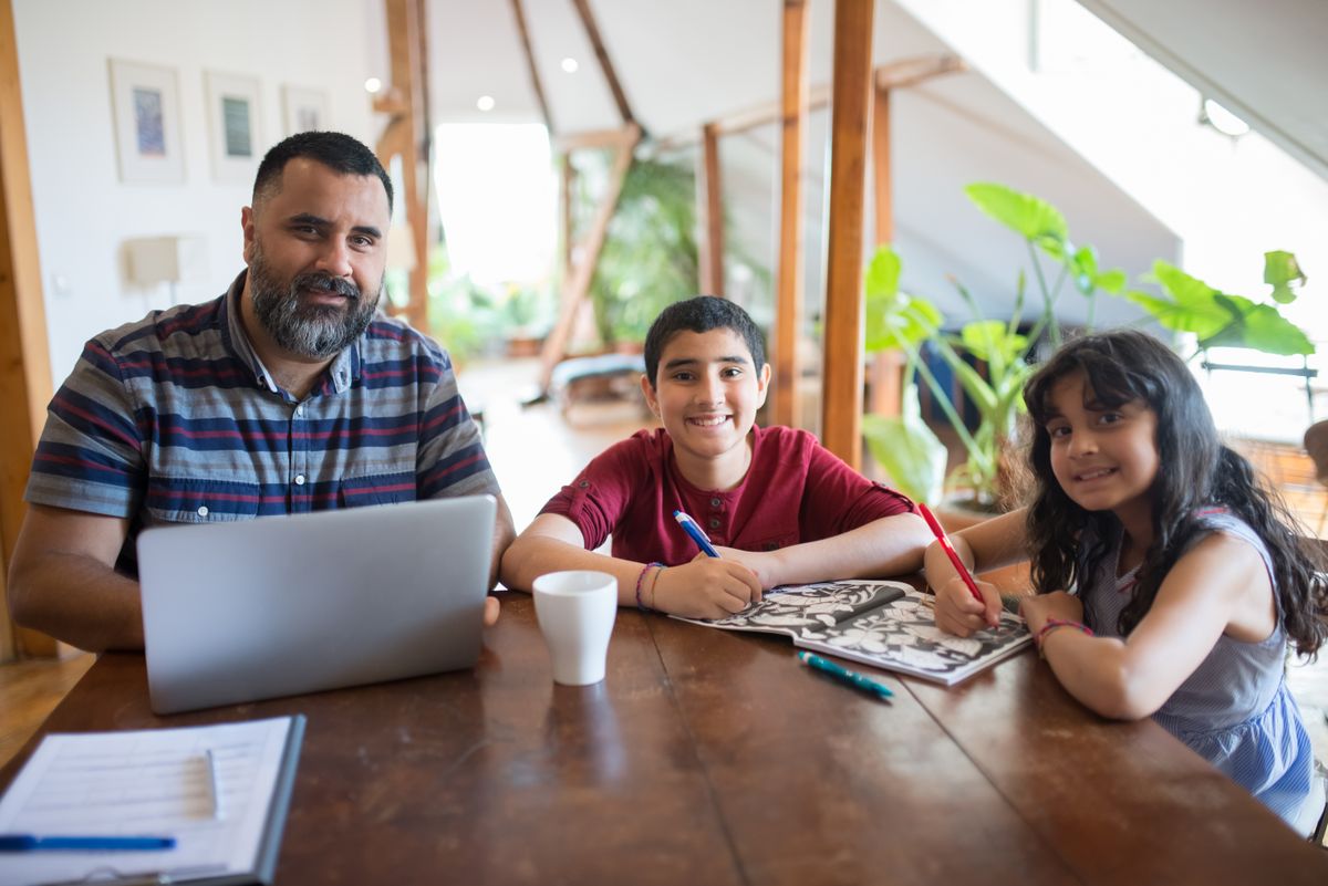 Father smiling at his laptop beside his son and daughter as they draw together at the table