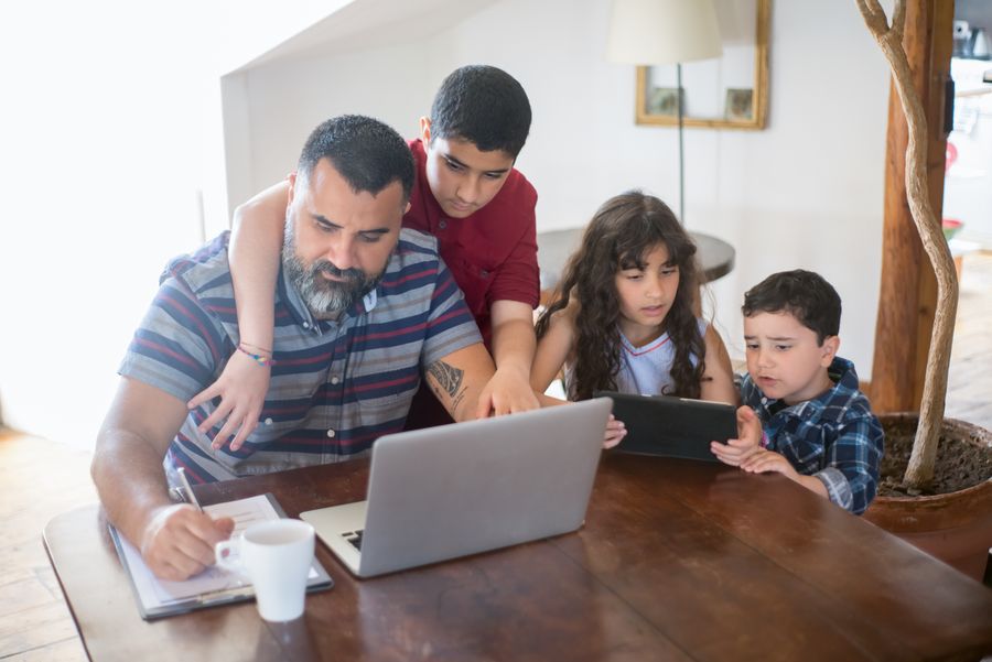 A father and three children gathered around a laptop and tablet at a home table