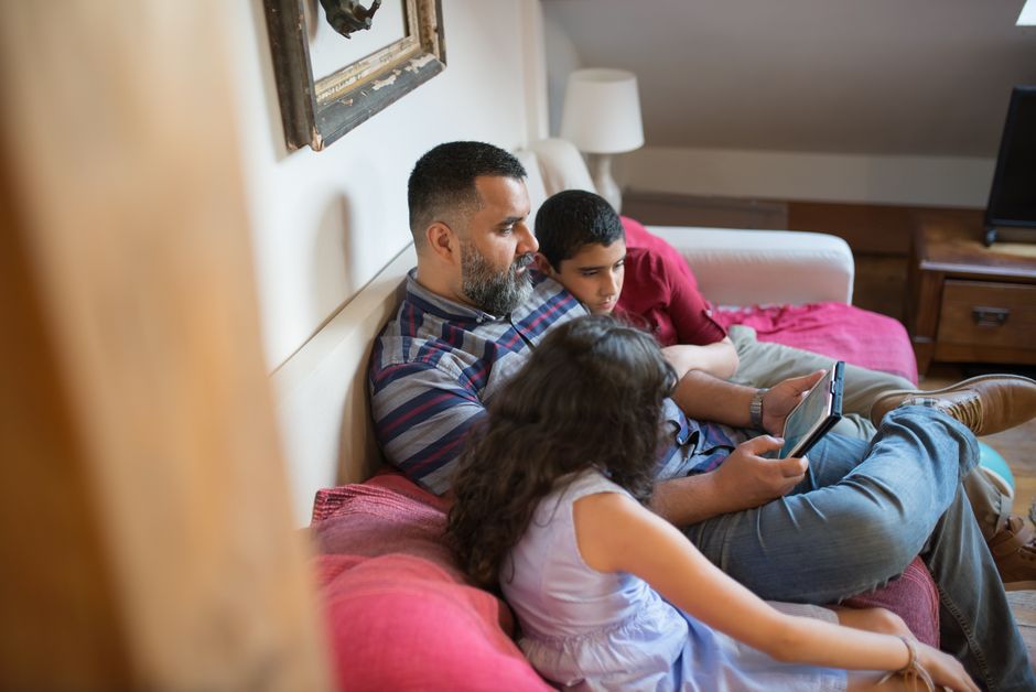 A father and his children relaxing on a couch using a digital tablet in a cozy home setting.