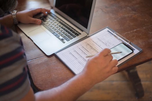 Close-up of a person using a laptop and reviewing documents with a smartphone on a wooden table.