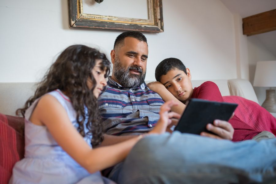 A parent and two children looking at a tablet together on a sofa at home