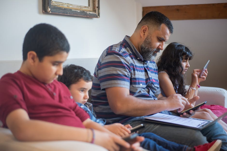 A father and three children engaging with technology while sitting on a sofa indoors.