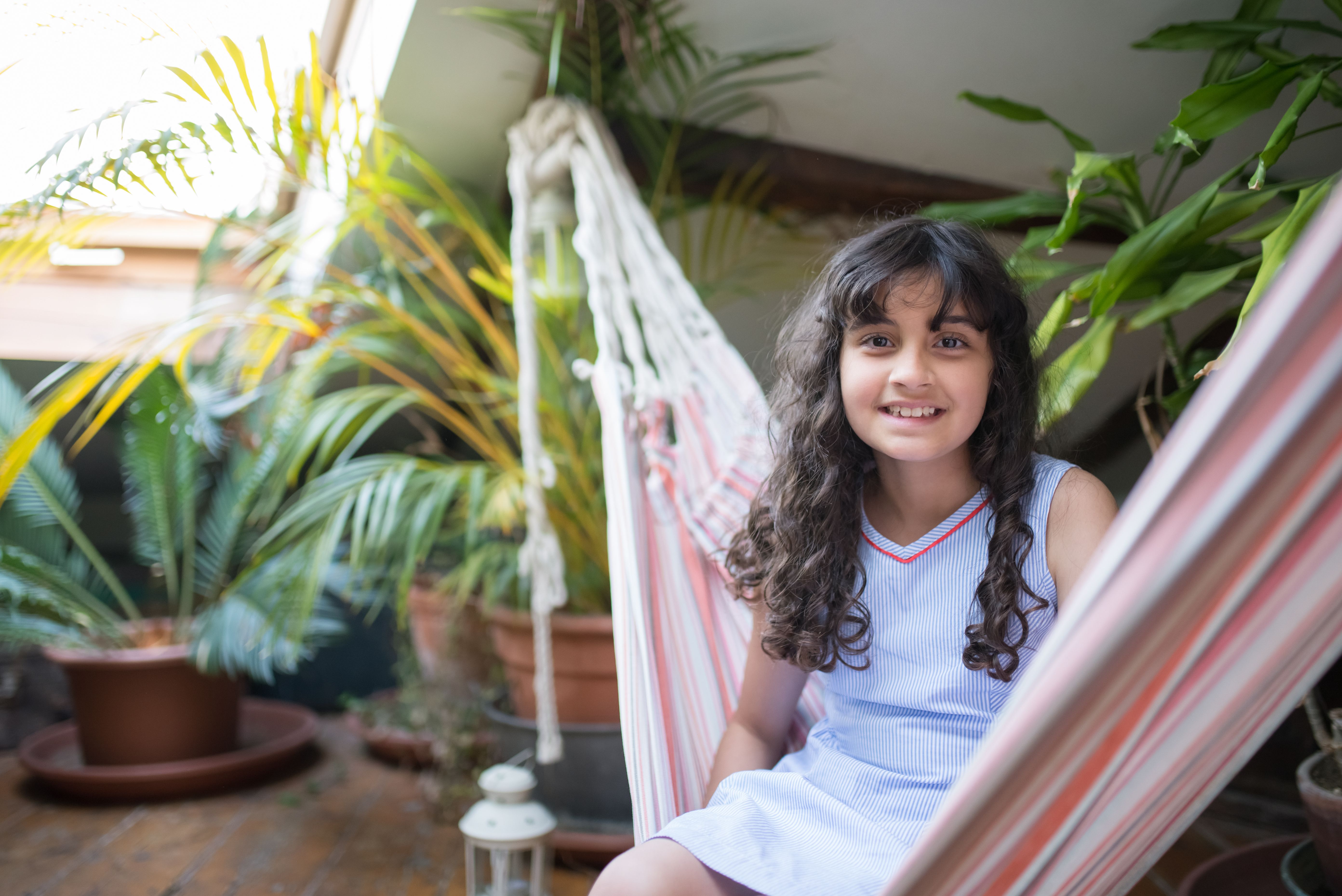 Young girl smiling while sitting in a hammock surrounded by plants indoors.