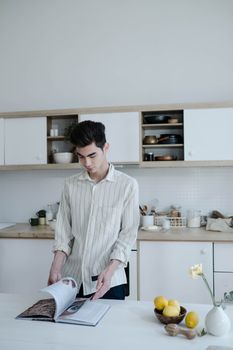 A young man in a striped shirt reading a cookbook in a modern minimalist kitchen.