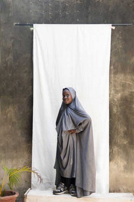 Portrait of a young Muslim girl in a modest outfit posing outdoors against a white backdrop.