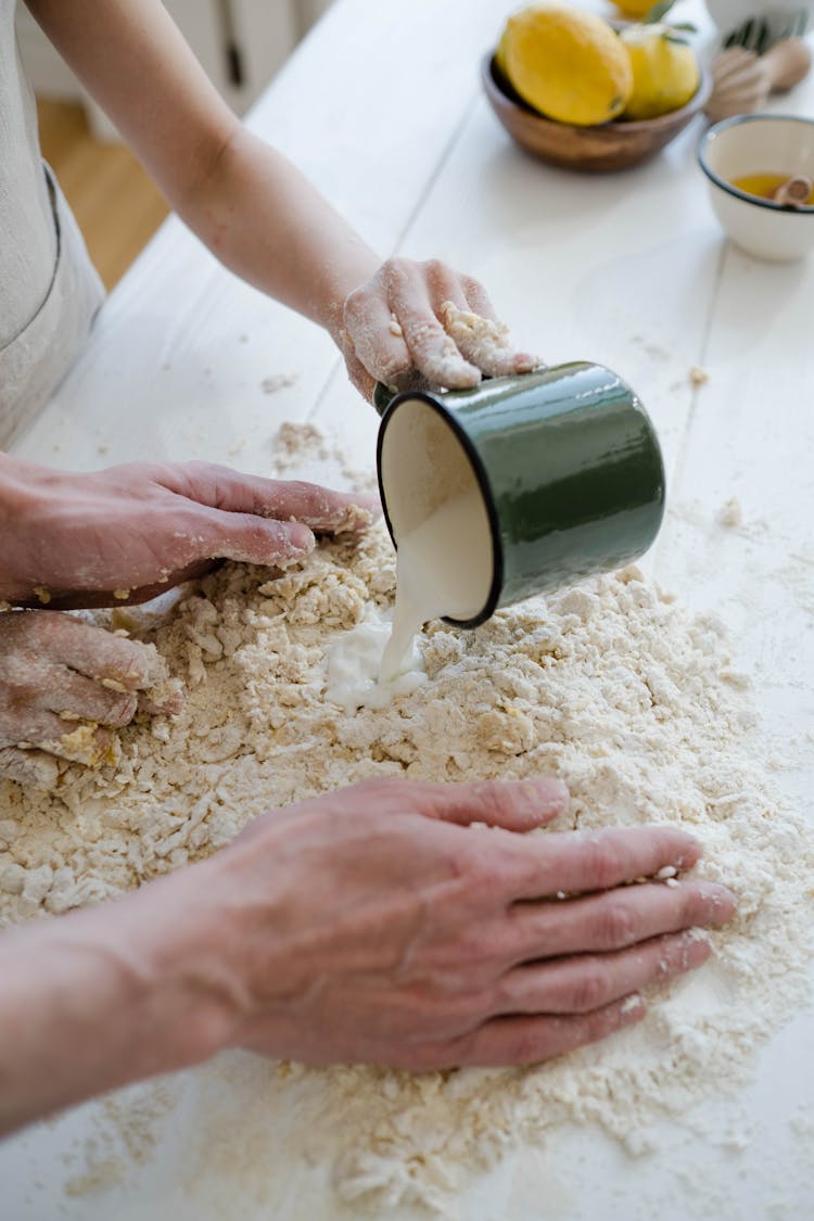 Hands Of Persons Mixing Milk And Flour On A White Surface