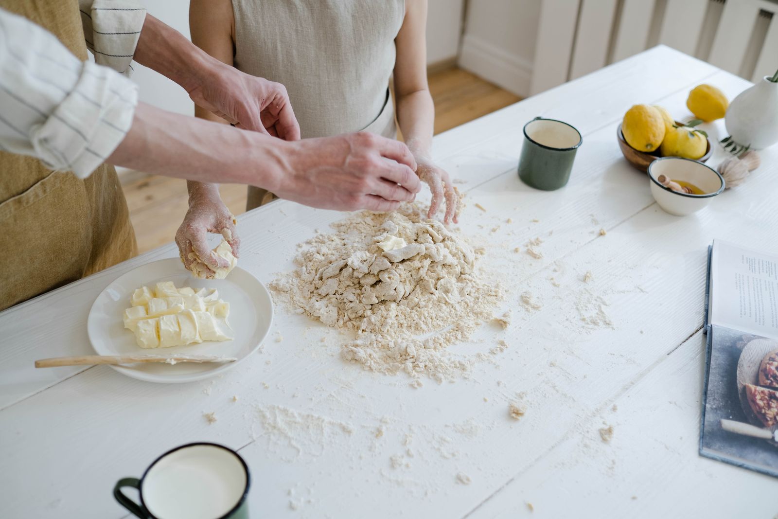 Two pairs of hands rubbing butter into flour on a white kitchen table beside lemons