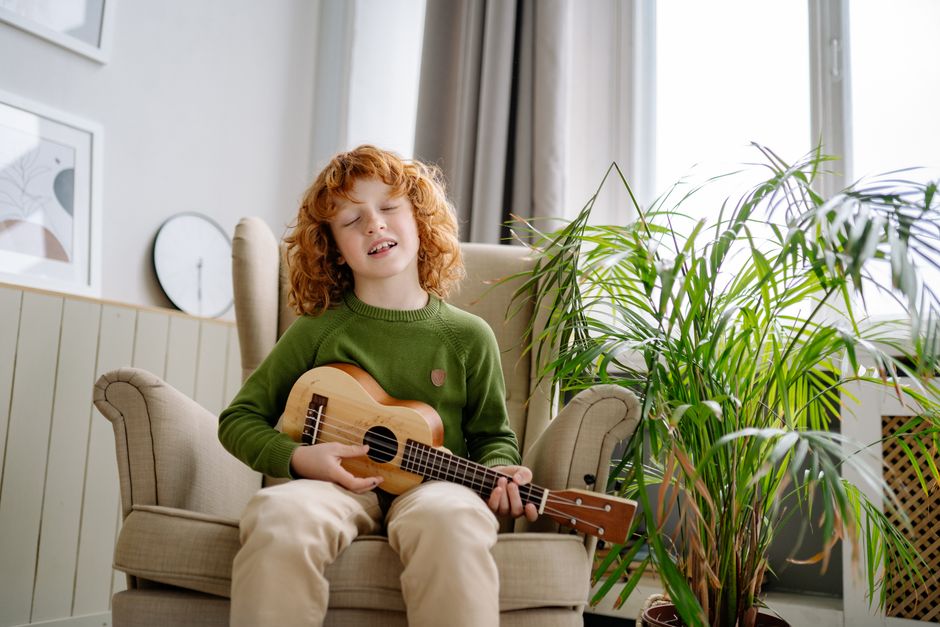 A red-haired child playing a ukulele while sitting on a chair indoors.