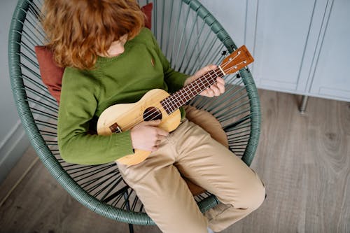 High-Angle Shot of a Redheaded Boy Playing Ukulele while Sitting on a Chair