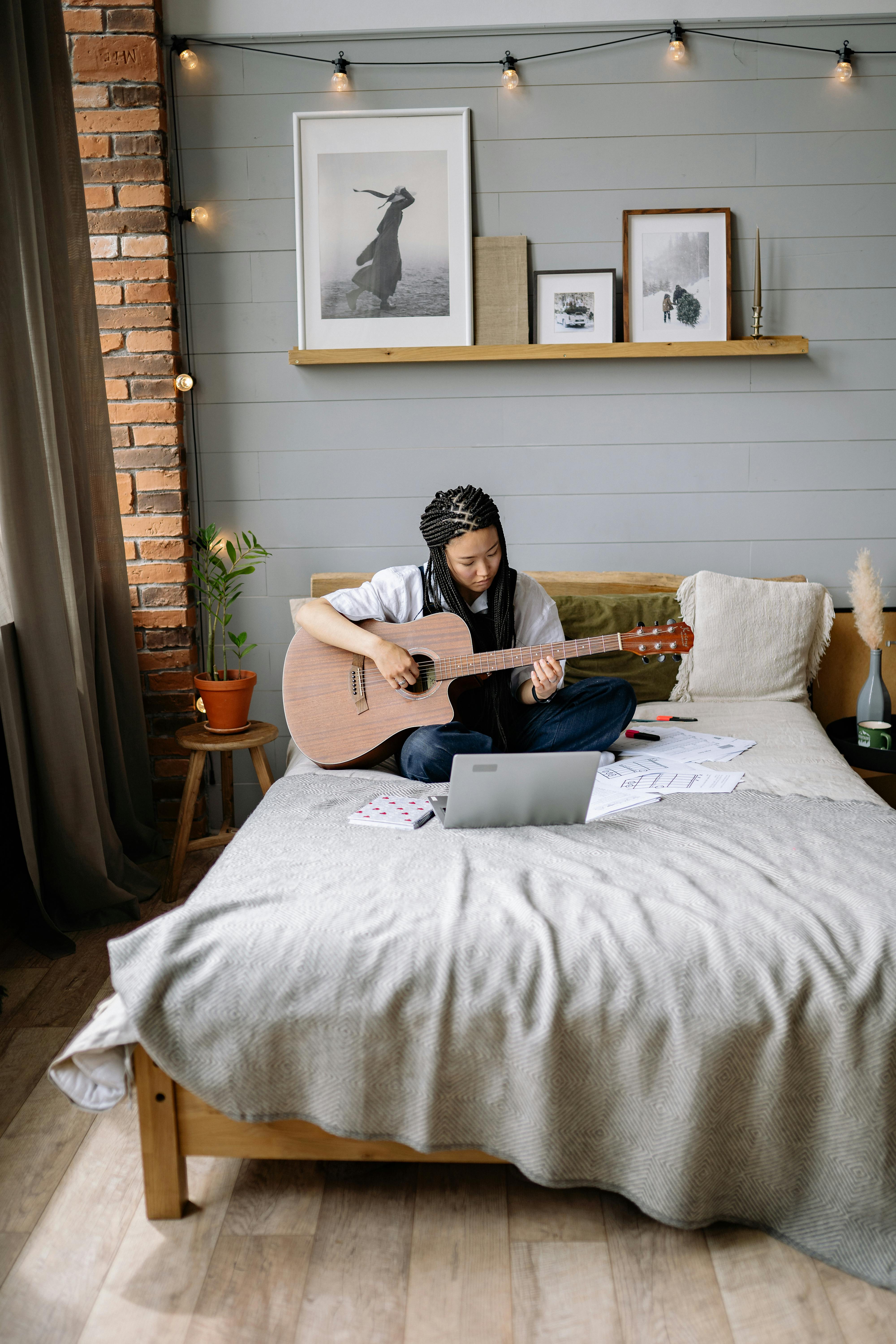 A woman enjoys playing acoustic guitar on her bed, surrounded by modern decor and a laptop for leisure and creativity.