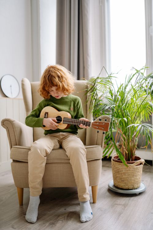 Free A redheaded child sitting on an armchair indoors, playing a ukulele beside a potted plant. Stock Photo
