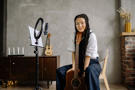 Asian woman with braids streaming music content at home, posing with an acoustic guitar, using a ring light.