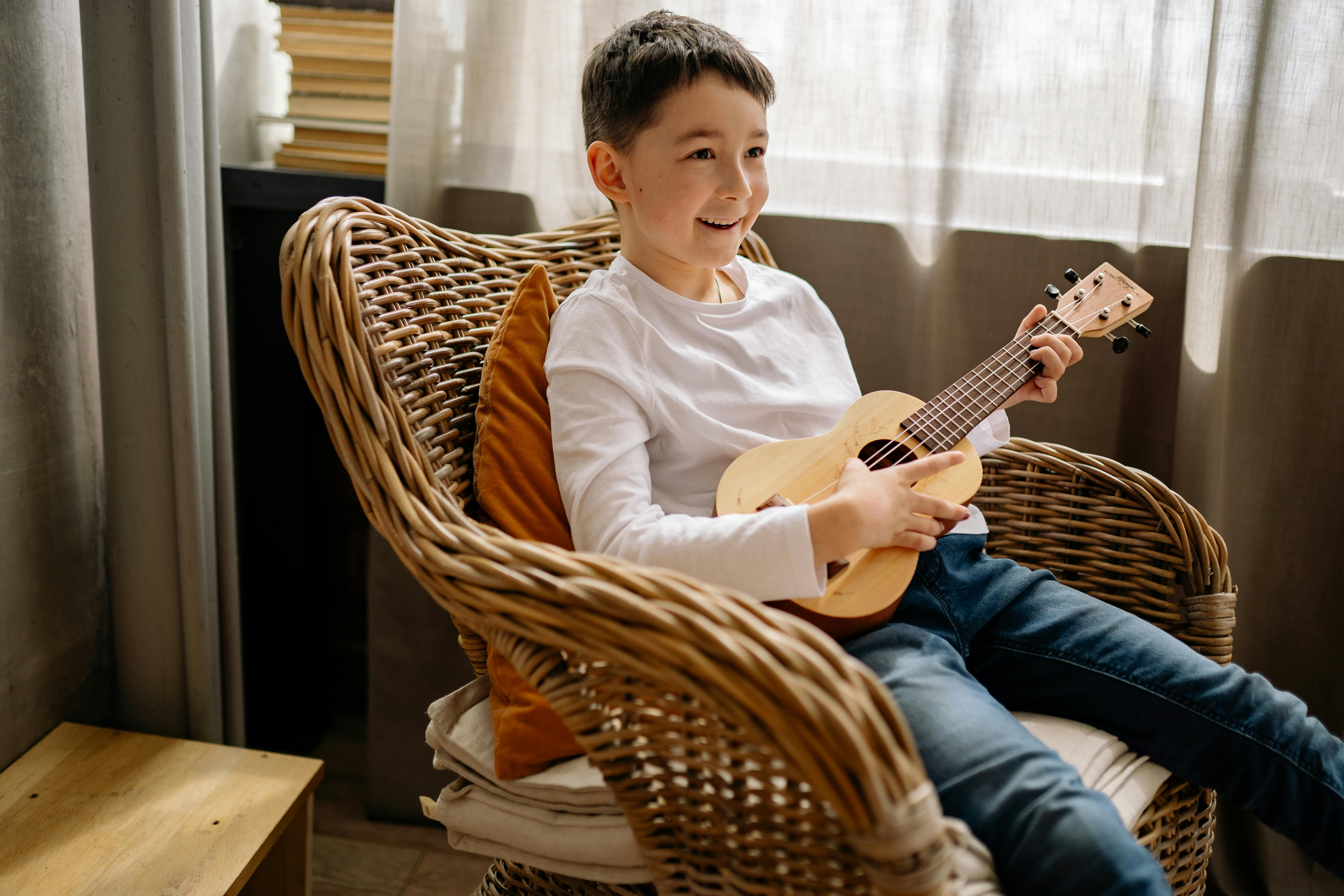 A cheerful child in a white shirt plays a ukulele, sitting on a woven chair indoors.