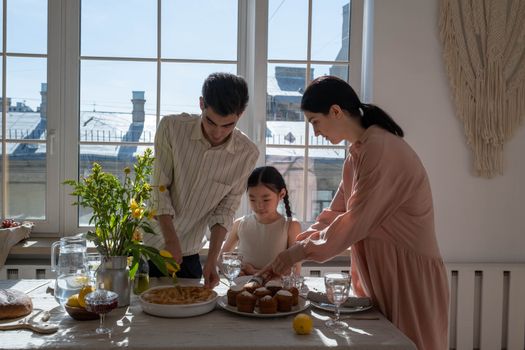 A family of three prepares a dinner table set with desserts and flowers, enjoying quality time indoors.