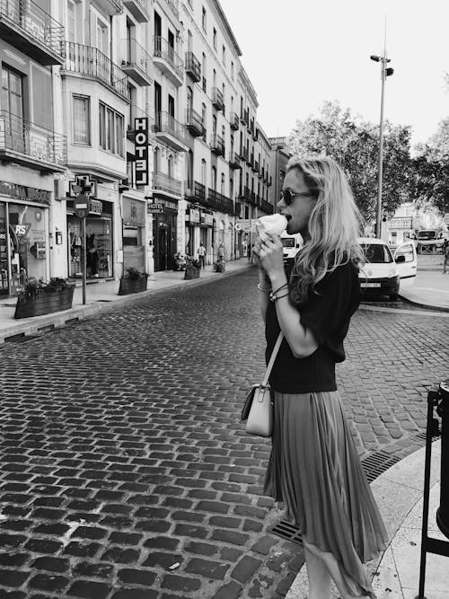 A Grayscale Photo of a Woman in Black Shirt Standing on the Street while Eating Food