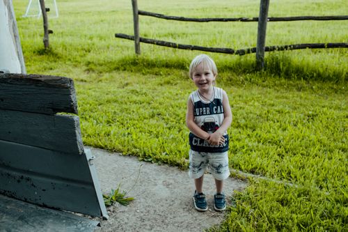 Free A smiling young boy stands in a grassy field with a rustic fence, exuding a happy and playful summer vibe. Stock Photo
