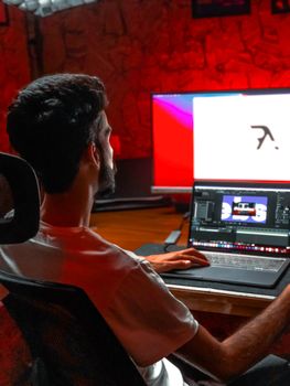 A man with a beard working on a laptop in a dimly lit room, focusing on screen.