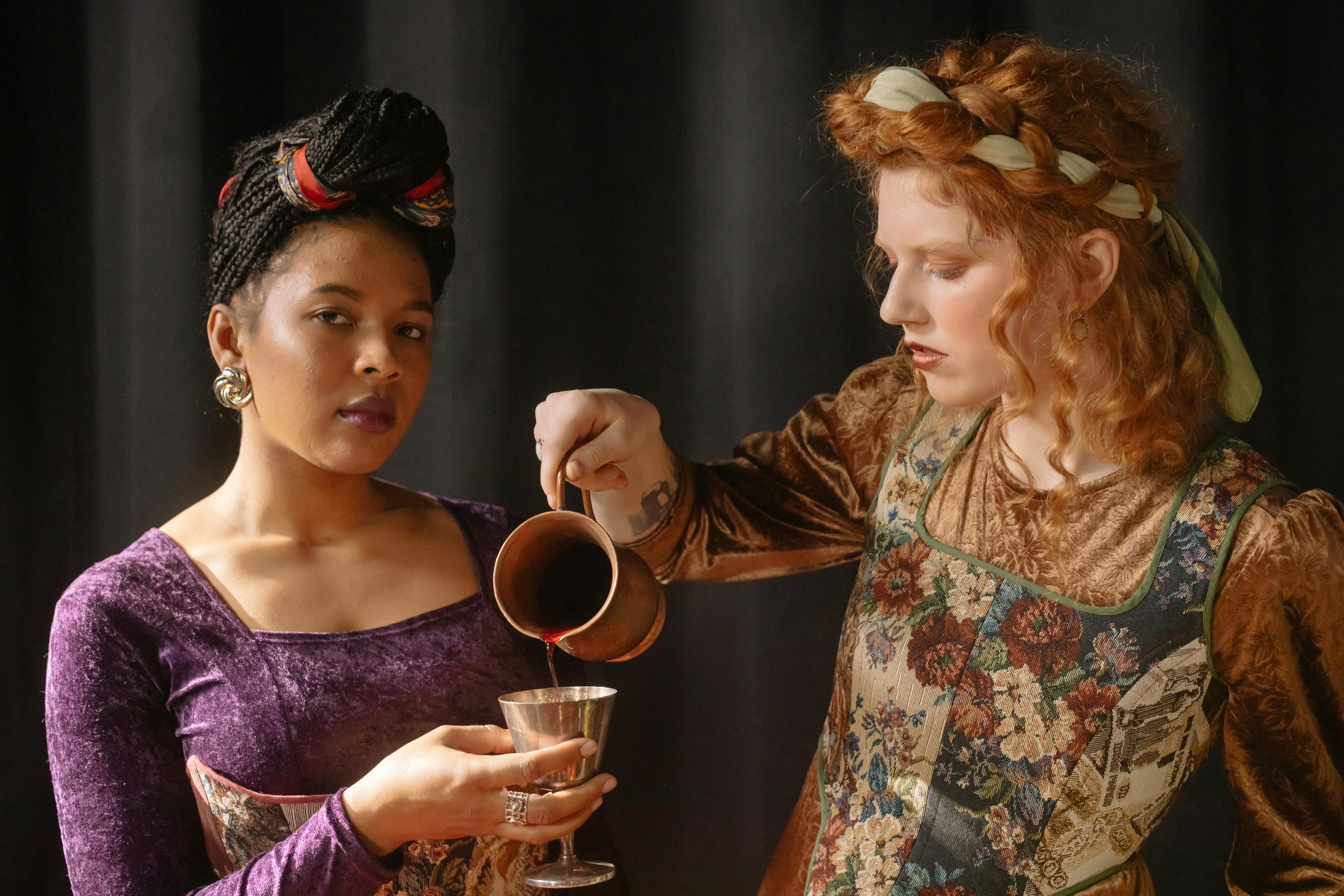 Two women in Renaissance-style attire pour wine from a metal cup.