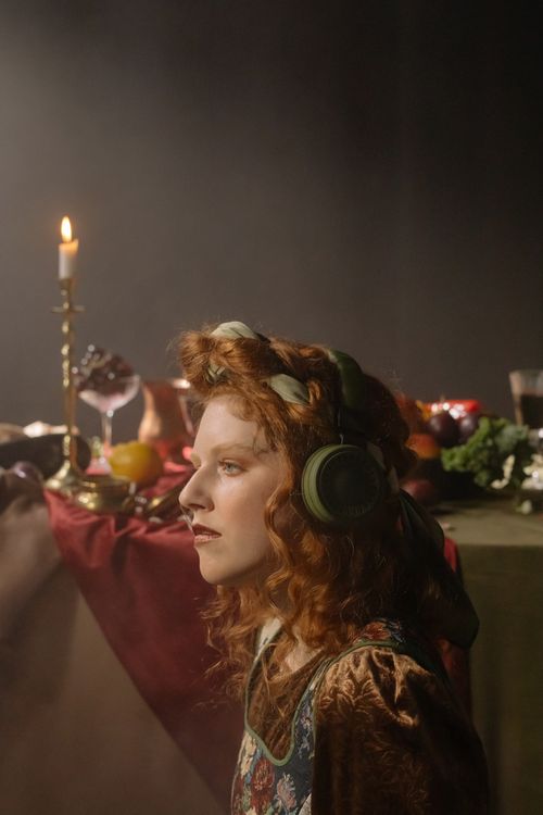 Free Red-haired woman in profile wearing headphones, seated in a vintage setting with a candlelit table. Stock Photo