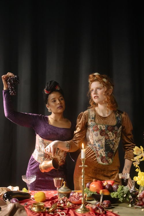 Free Two women in vintage clothing enjoy a candlelit medieval-inspired feast with grapes and pomegranates. Stock Photo