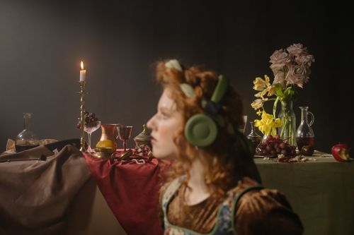 Free A woman in headphones sits near a classic still life table with candles, fruits, and flowers. Stock Photo