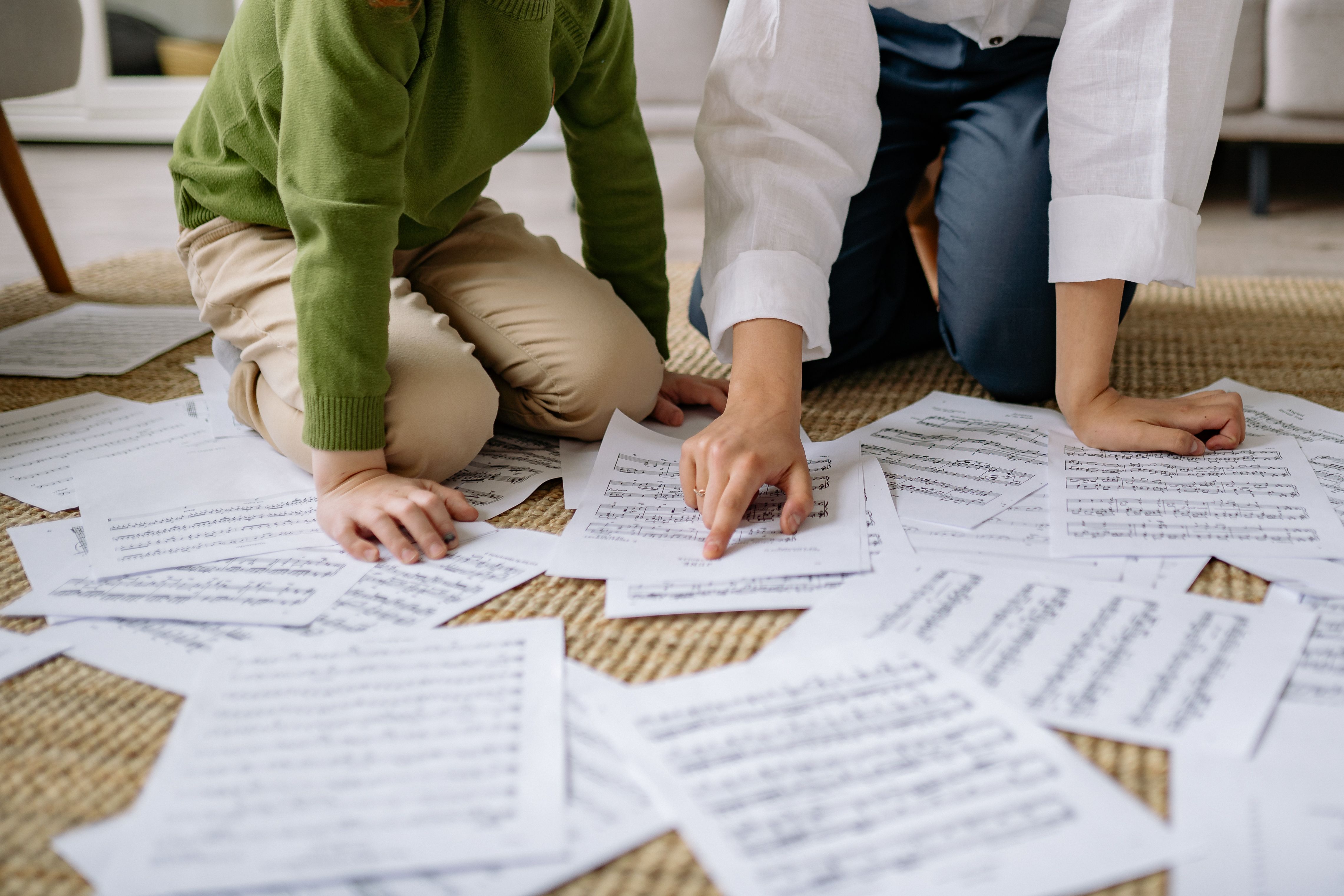A child and adult kneeling indoors, analyzing scattered music sheets on the floor.