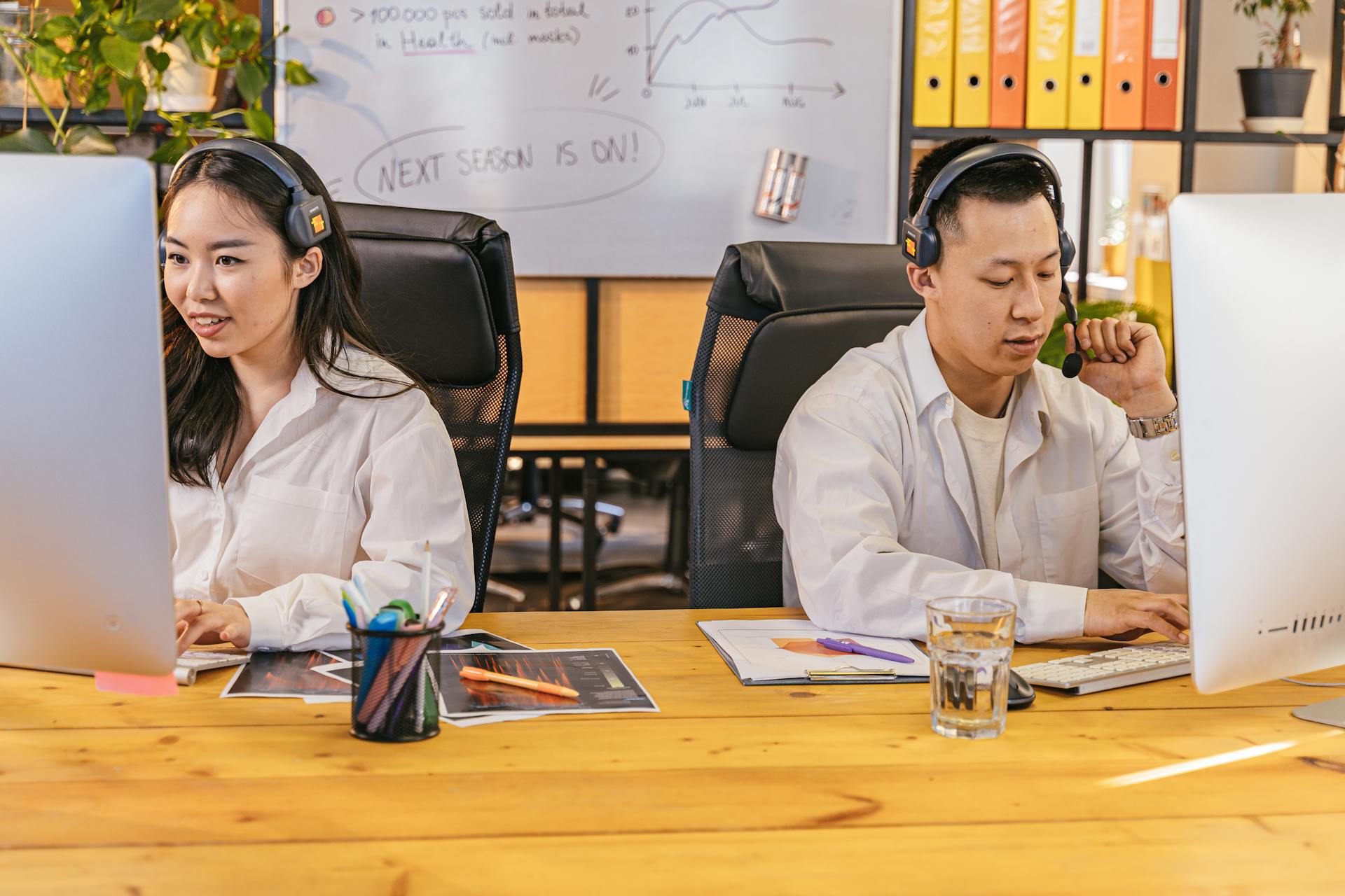 Two professionals at desks using computers and headsets in a modern office setting.