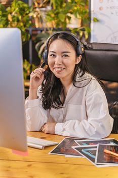 Asian woman smiling while working at a desk with a headset and computer indoors.
