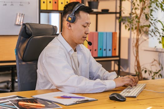 Asian man wearing headset working on a computer in a modern office environment.