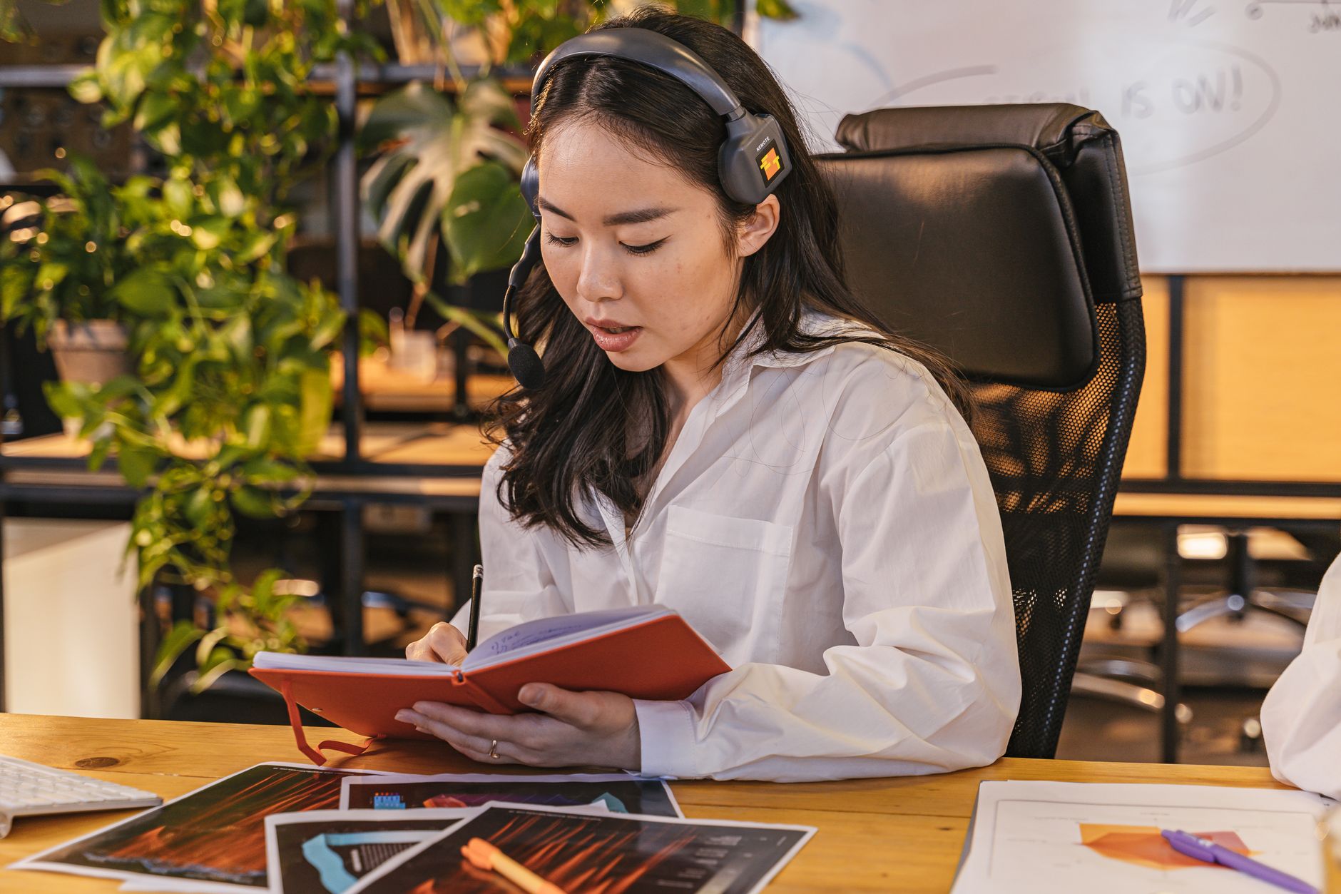 Professional Asian woman with headset taking notes at a desk in an office setting.