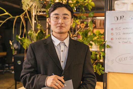 A confident young businessman in a suit and eyeglasses standing in a modern office.