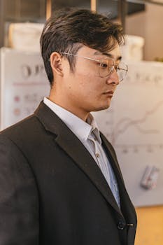 Side profile of a man in a black suit with eyeglasses, focused on data analysis in an office.