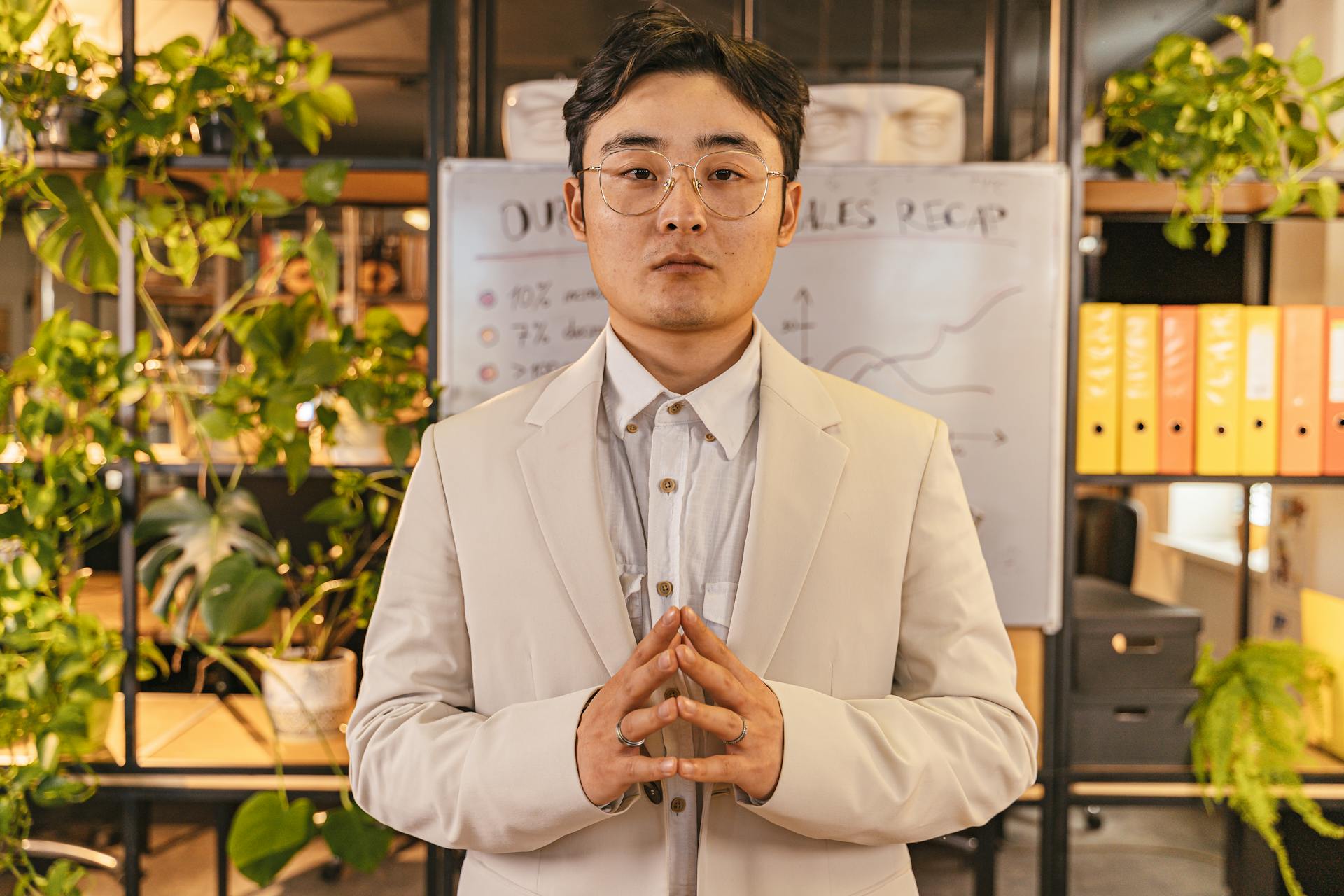 Young professional standing confidently in an office with greenery and a sales recap board.