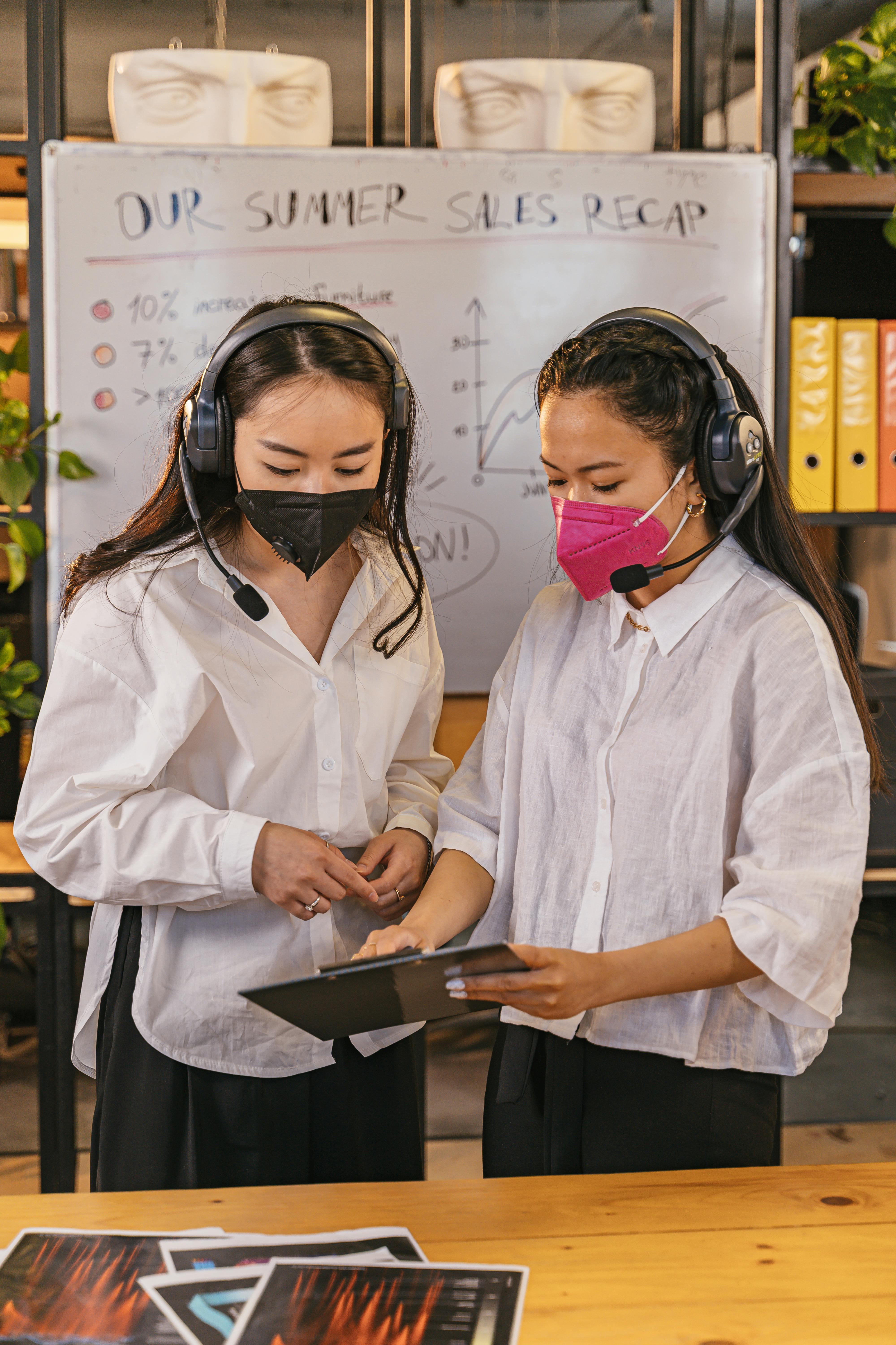 Two businesswomen wearing masks and headsets in a collaborative office meeting.