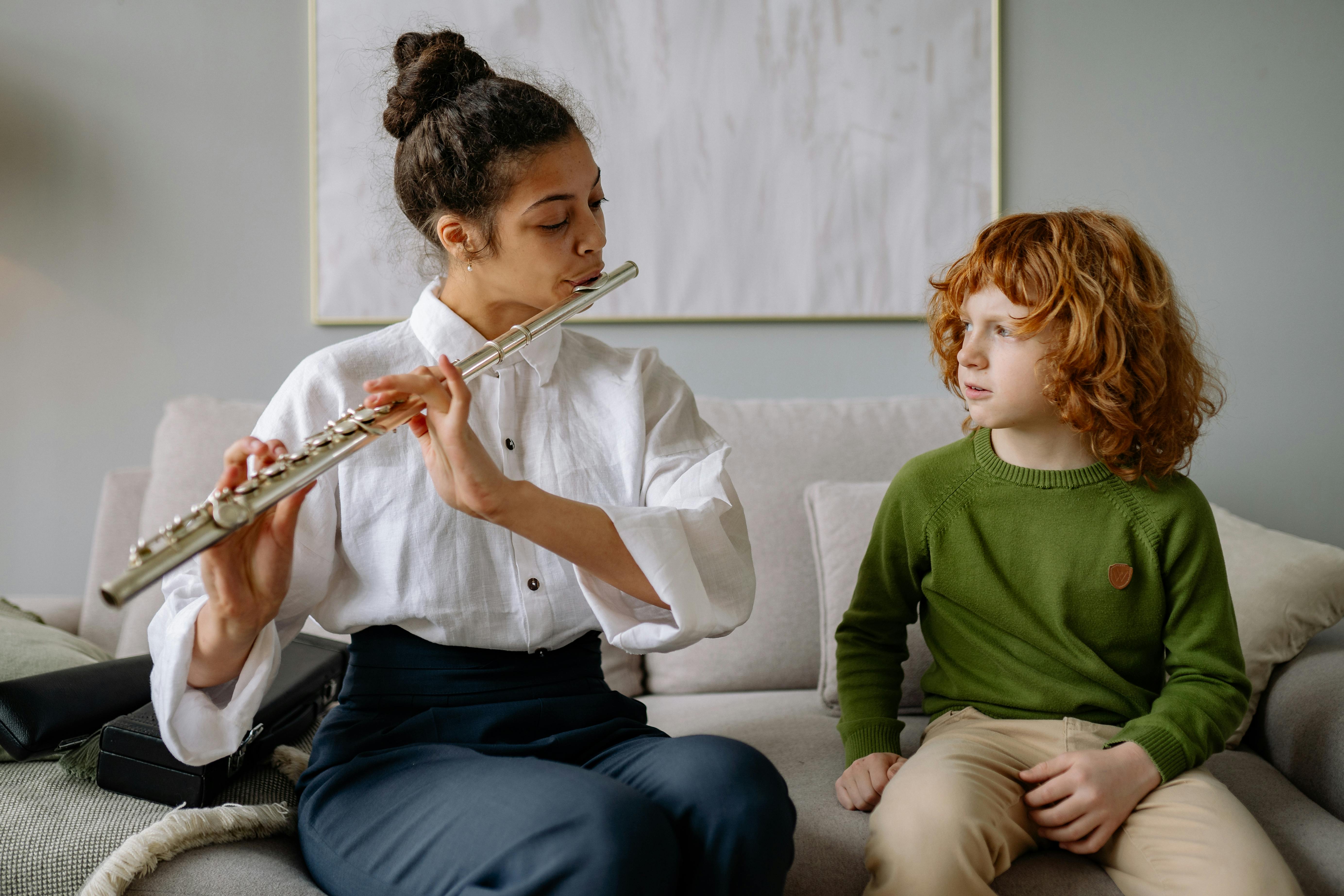 A woman teaching a child to play the flute on a sofa during a music lesson indoors.