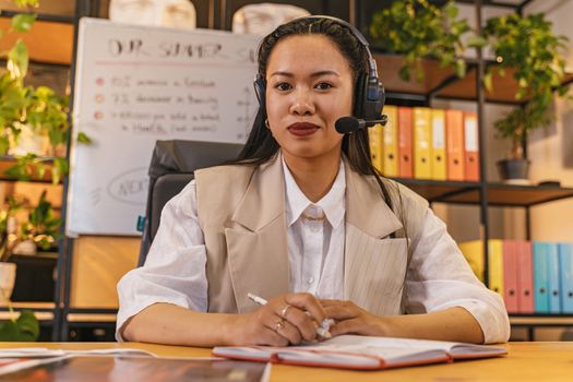 Asian woman in headset smiling while working in a modern office environment.